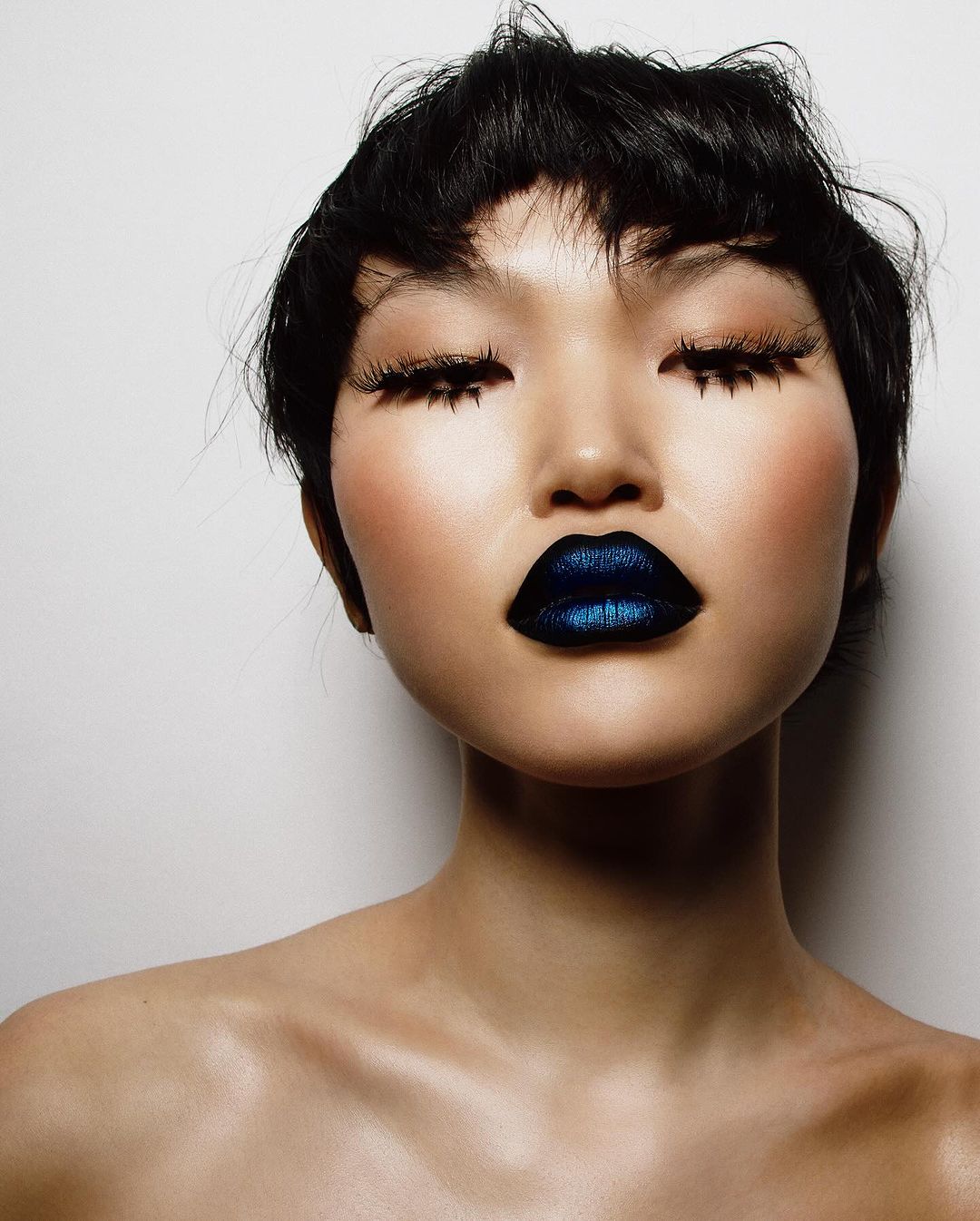 Close-up portrait of a bare-shouldered Asian woman with dark hair, closed eyes, dramatic long eyelashes, and shimmering blue lipstick against white.