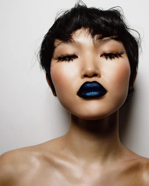 Close-up portrait of a bare-shouldered Asian woman with dark hair, closed eyes, dramatic long eyelashes, and shimmering blue lipstick against white.