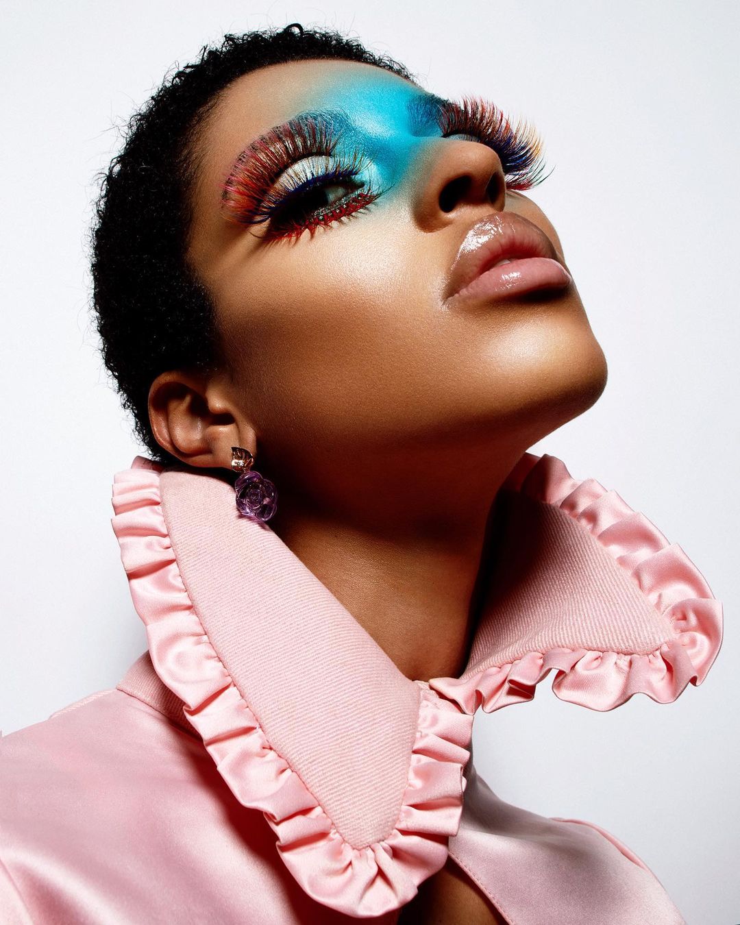 Glamorous beauty shot of a short-haired model in a pink ruffled collar with colorful eye makeup, long lashes, and glossy lips on a white backdrop.