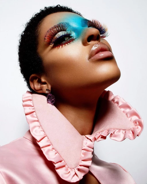 Glamorous beauty shot of a short-haired model in a pink ruffled collar with colorful eye makeup, long lashes, and glossy lips on a white backdrop.