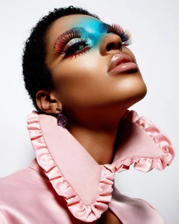 Glamorous beauty shot of a short-haired model in a pink ruffled collar with colorful eye makeup, long lashes, and glossy lips on a white backdrop.