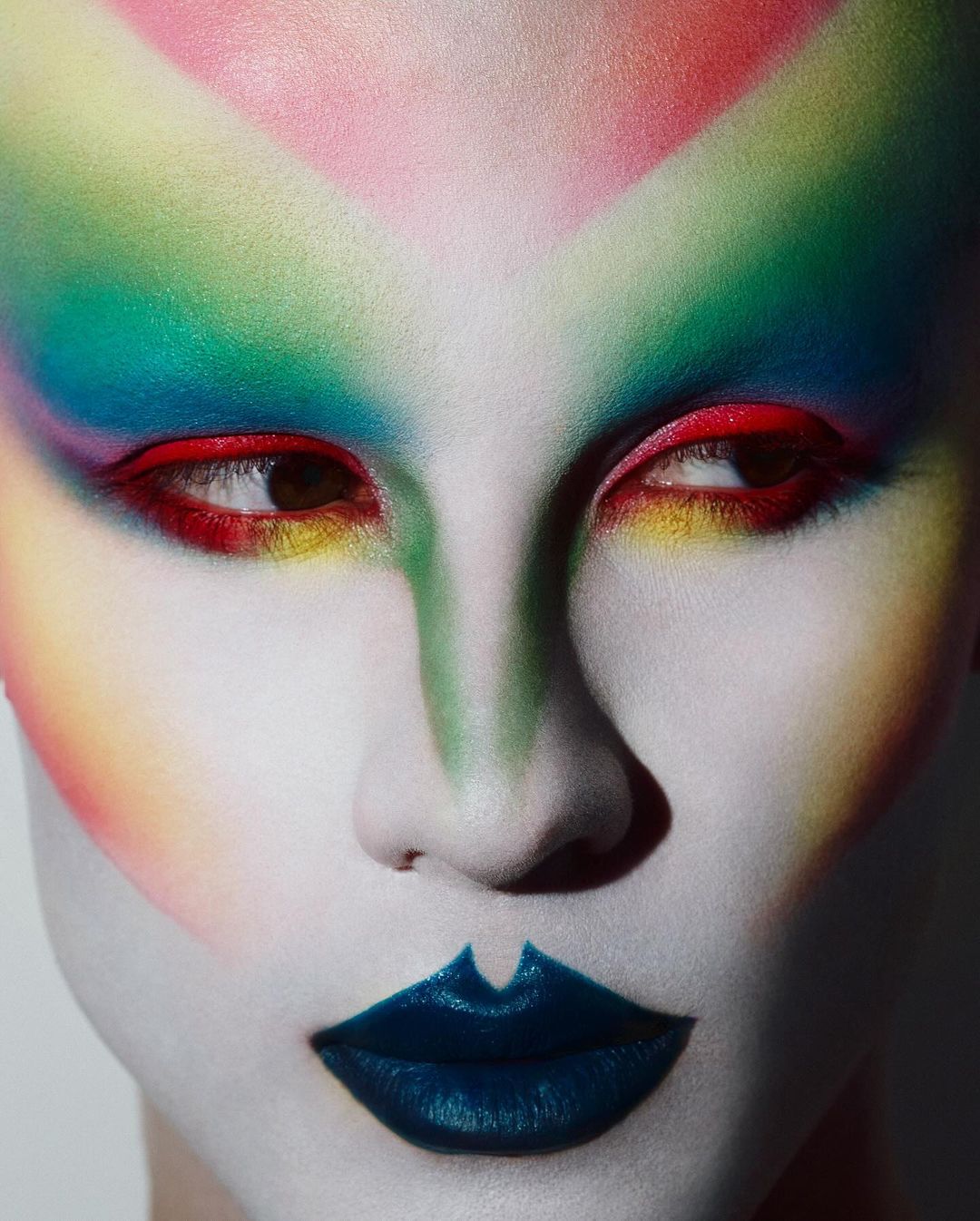 Dramatic beauty shot of a person with face paint in rainbow colors, white foundation, and dark blue lips, close-up of their face looking to the side.