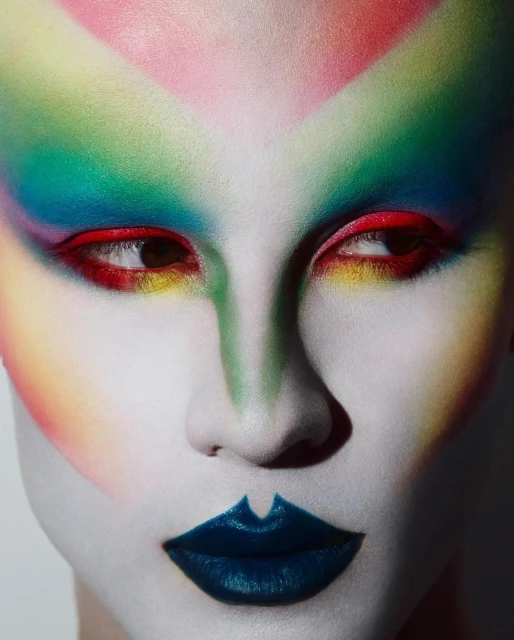 Dramatic beauty shot of a person with face paint in rainbow colors, white foundation, and dark blue lips, close-up of their face looking to the side.