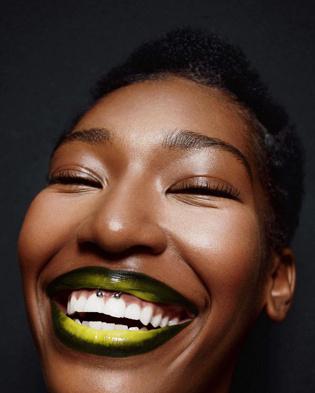 Close-up of a smiling Black woman with yellow and green lipstick, visible teeth, and a lip piercing against a dark background.