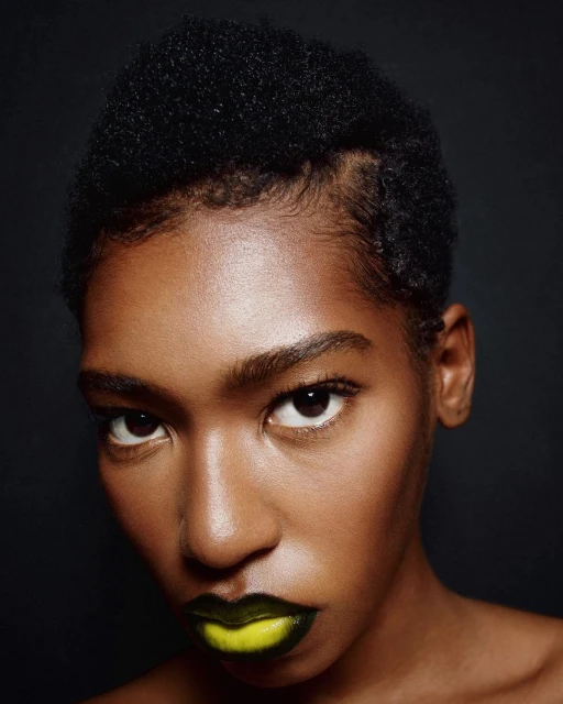 Headshot of a young Black woman with short dark hair, wearing bright yellow lipstick, against a dark background in a beauty style.