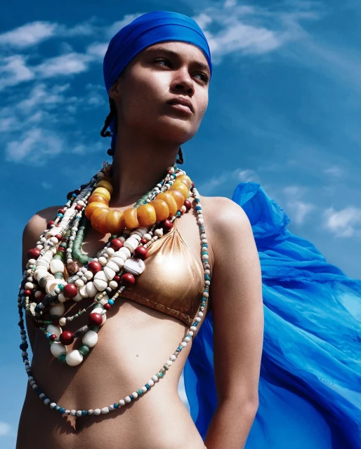 Woman in headscarf wearing layered necklaces, stands against a blue sky in a gold bikini top, creating a fashion-forward visual.