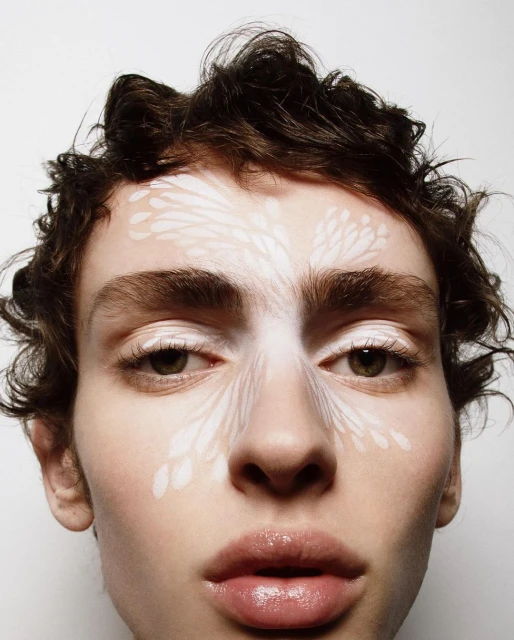 Close-up portrait of a fair-skinned person with wavy brown hair and pale-toned makeup design on their forehead and between their eyes.