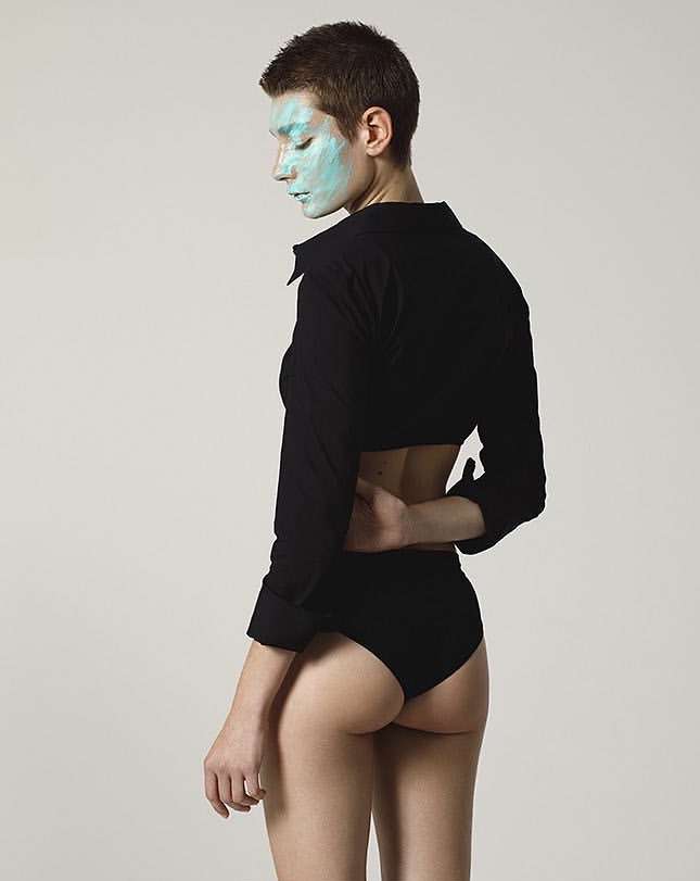 Young woman poses in black top and briefs with light blue facial mask, set against a neutral backdrop in a studio-style fashion shot.