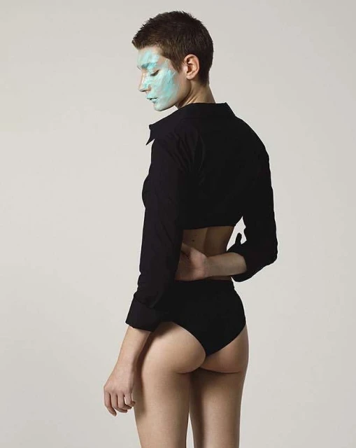 Young woman poses in black top and briefs with light blue facial mask, set against a neutral backdrop in a studio-style fashion shot.