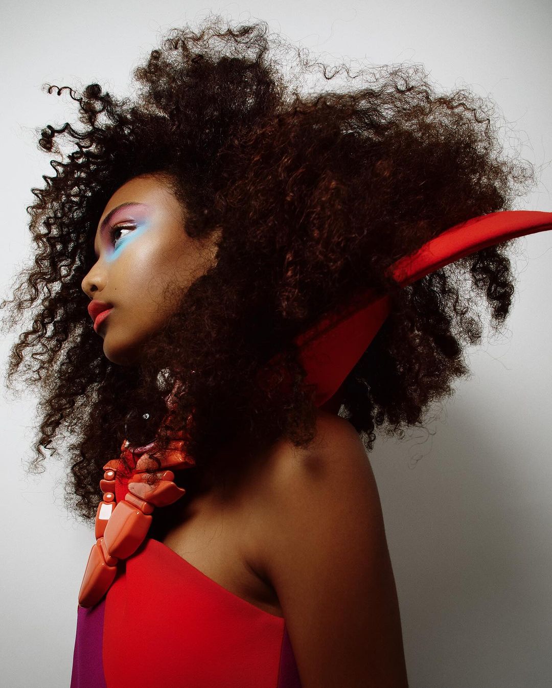 Close-up portrait of a Black woman with dramatic makeup, voluminous afro hairstyle, and a sculptural red necklace and headpiece against white.