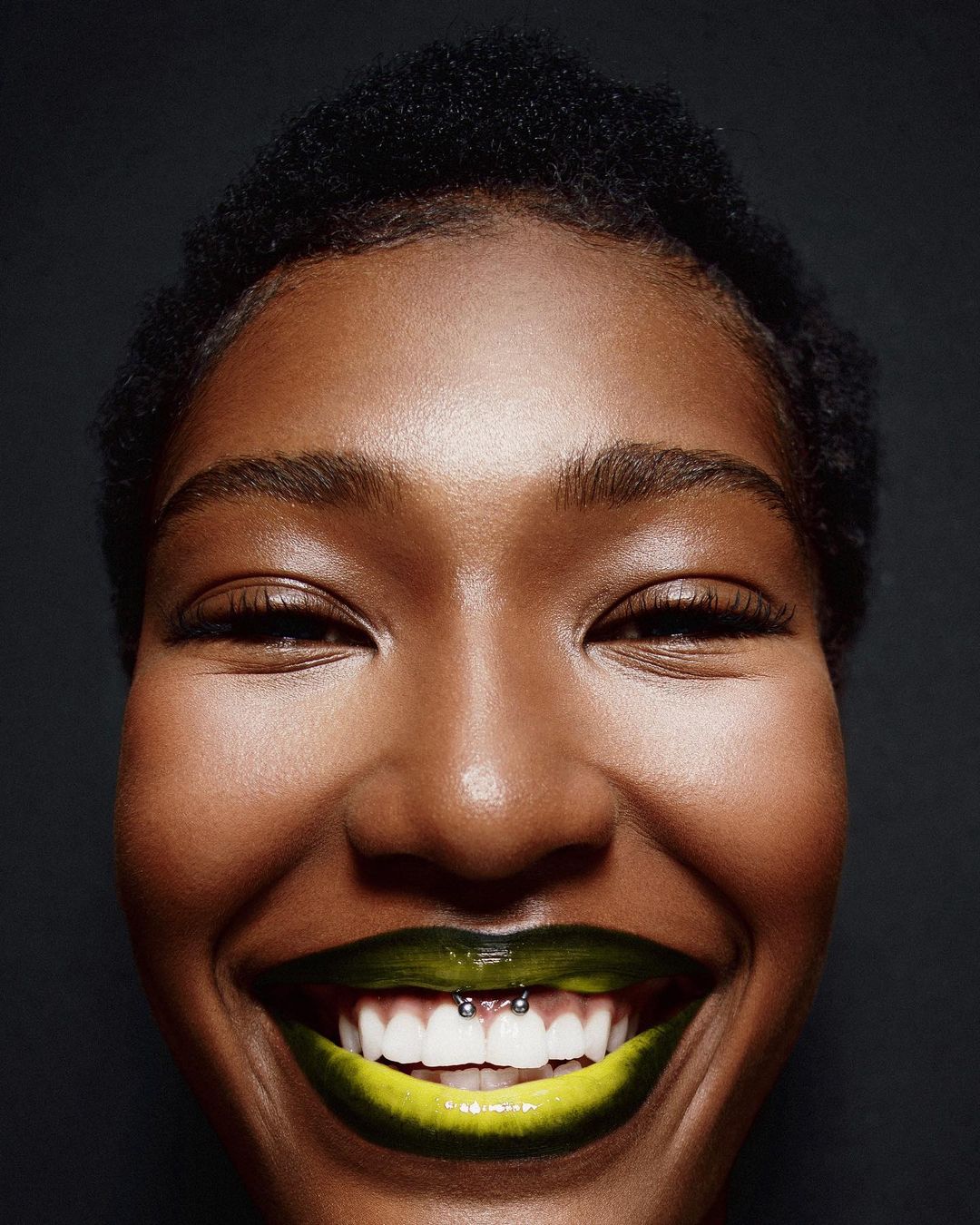Close-up portrait of a smiling African-American woman with a tooth piercing and vibrant yellow-green lipstick against a dark gray background.