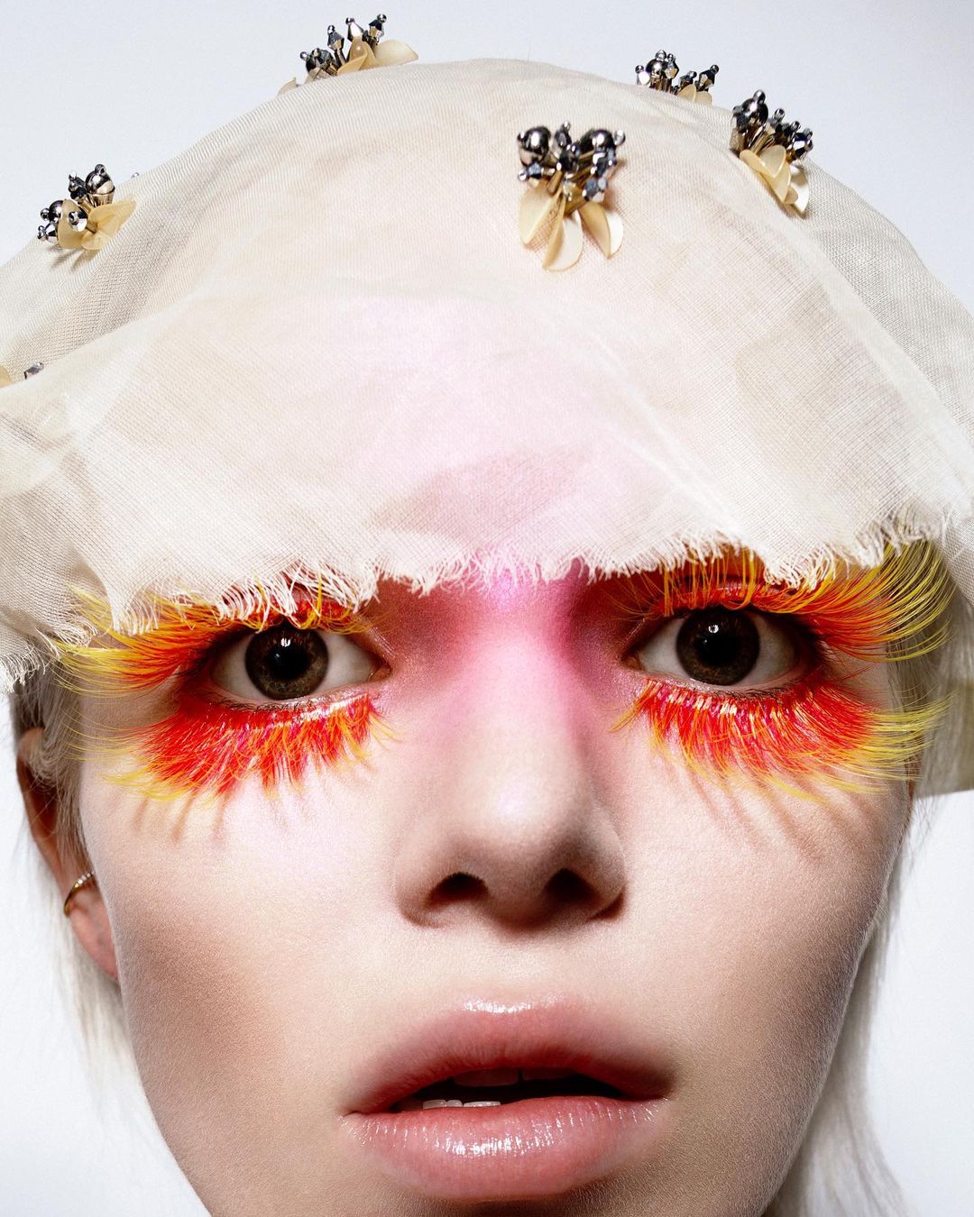 Close-up portrait of a fair-skinned person with decorative eyelashes, a sheer head covering, and pink eyeshadow, against a white backdrop.