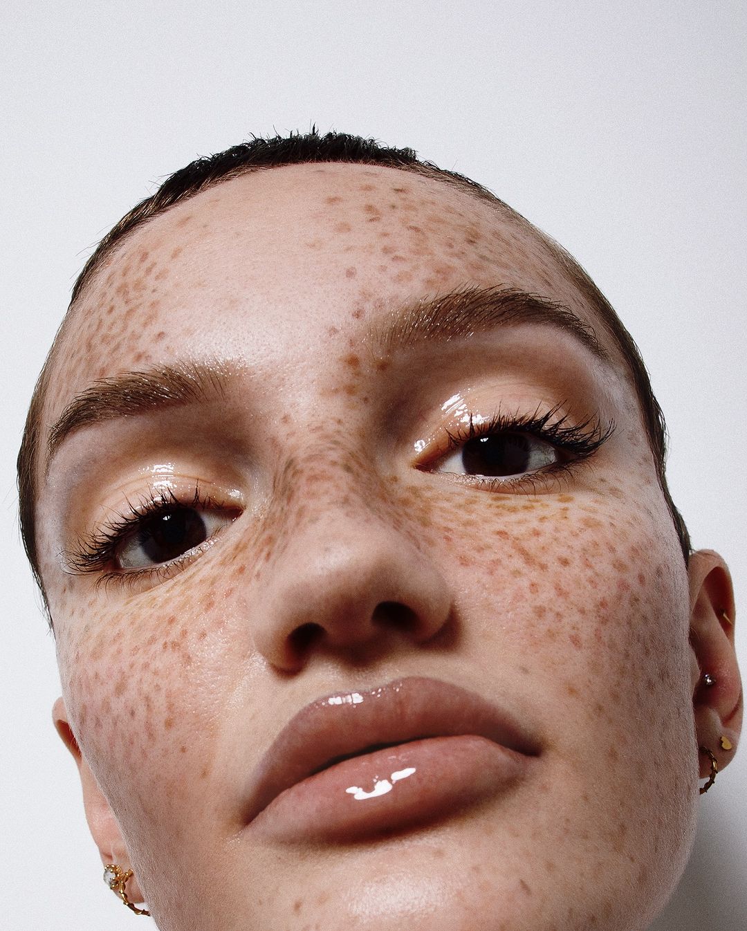 Close-up portrait of a woman with freckles and glossy makeup on her face, taken in a brightly lit photo studio against a white background.