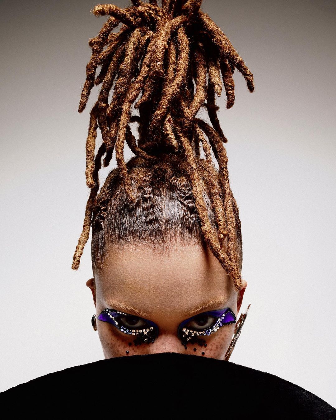 Eye-level close-up of a woman's face with dreadlocks, vibrant purple and silver eye makeup, and freckles, against a gradient grey background.