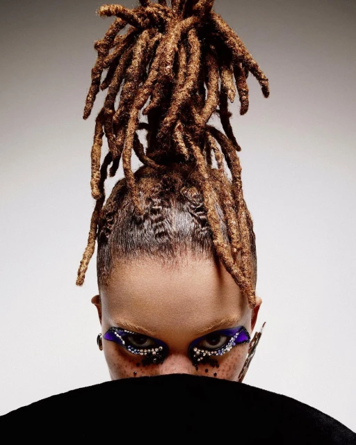 Eye-level close-up of a woman's face with dreadlocks, vibrant purple and silver eye makeup, and freckles, against a gradient grey background.