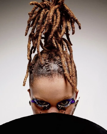 Eye-level close-up of a woman's face with dreadlocks, vibrant purple and silver eye makeup, and freckles, against a gradient grey background.