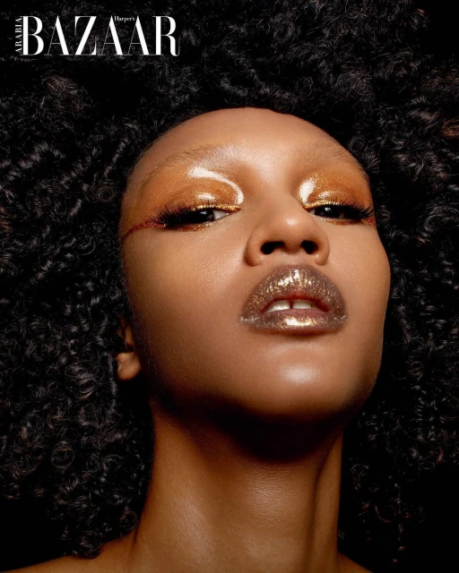 Beauty shot of a woman with dark curly hair, wearing metallic gold makeup, facing the camera with a low angle, against a dark backdrop.