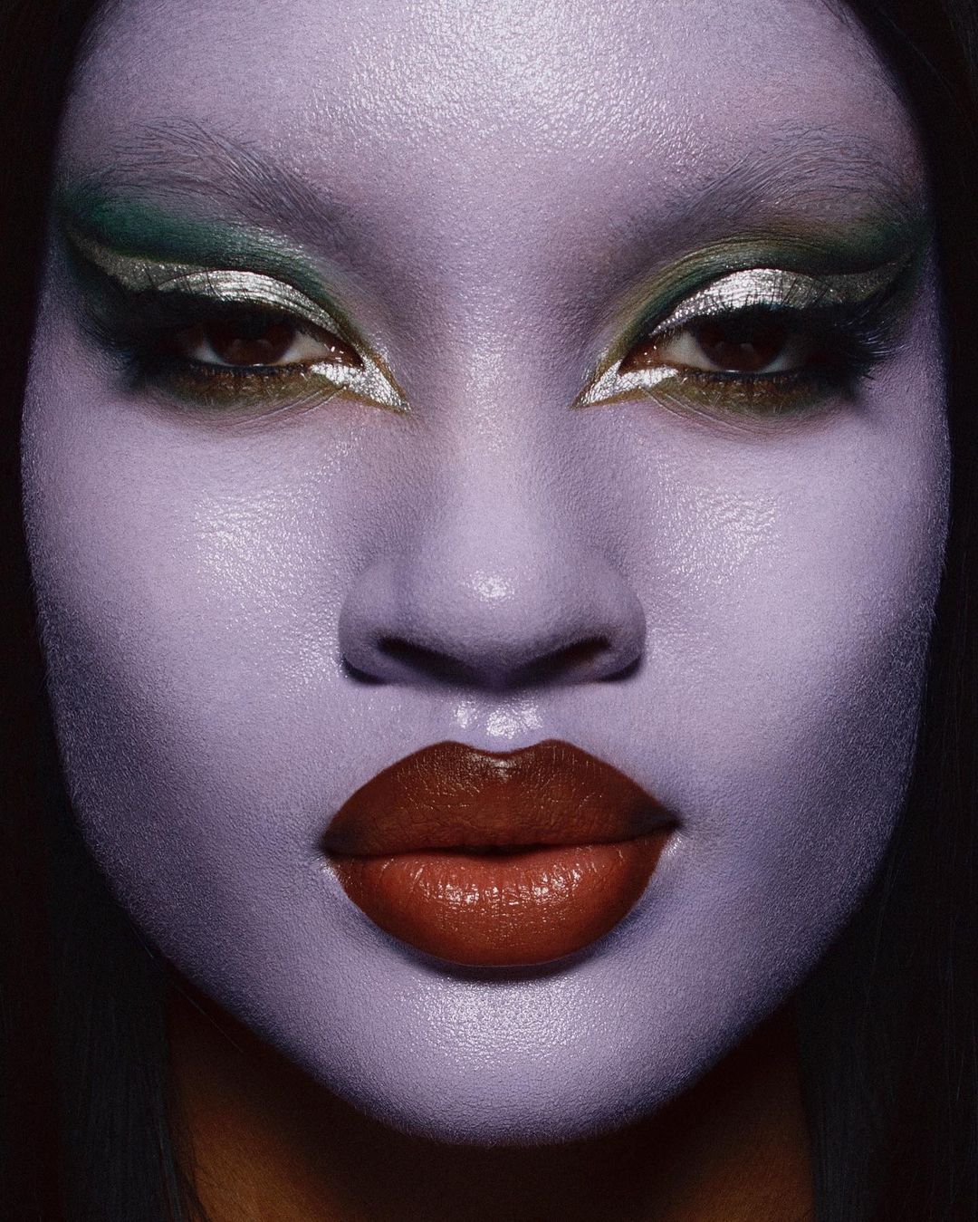 Close-up portrait of a woman with lavender face paint and creative silver and green eye makeup looking directly at the camera.