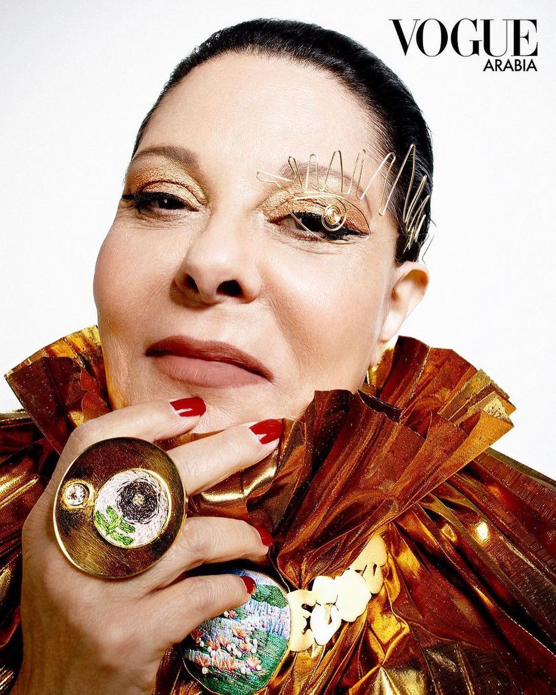 Portrait of an adult female wearing sculptural gold eye makeup, a large gold ring, and a shiny gold ruffled collar against a white background.