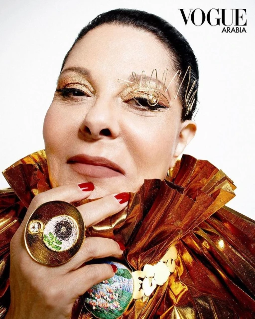 Portrait of an adult female wearing sculptural gold eye makeup, a large gold ring, and a shiny gold ruffled collar against a white background.