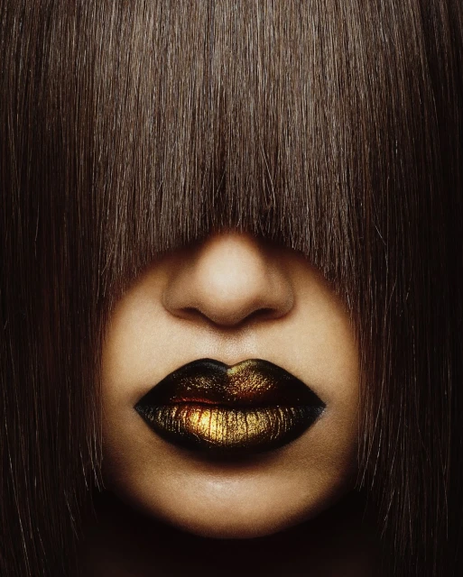 Dramatic close-up of a person's face partially obscured by dark brown hair, featuring gold and black lipstick.