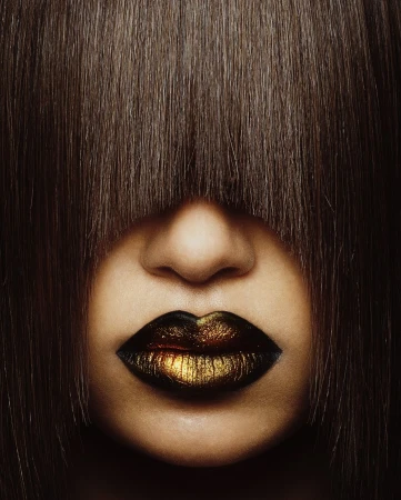 Dramatic close-up of a person's face partially obscured by dark brown hair, featuring gold and black lipstick.