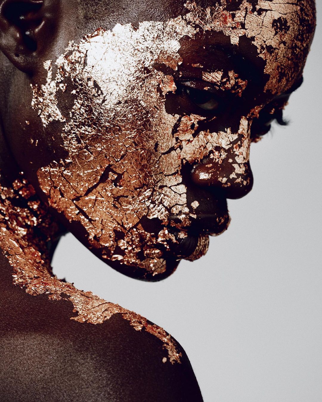 Portrait of a dark-skinned person with gold leaf applied to their face and upper chest, posing against a light background in studio lighting.
