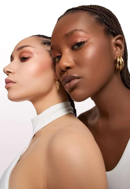 Two women pose in a beauty shot with neutral makeup and braided hairstyles, highlighted by a bright white background and gold earrings.