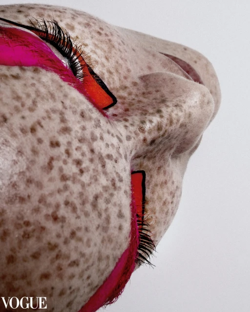 Extreme close-up of a person's face with pink and red geometric makeup, pale skin with freckles, and dark eyelashes against white.