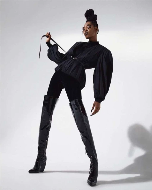 A stylish Black woman poses indoors wearing a black top, black leggings, and high patent leather boots in a fashion-forward shot.