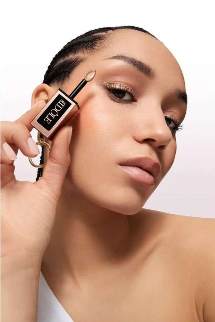 Close-up of a young woman with bronze eyeshadow holding a 'Idôle' product to her face, set against a bright white background.