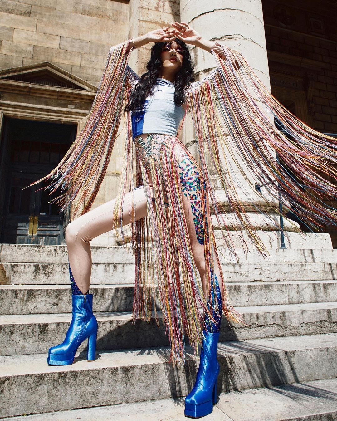A fashion shot of a woman in a colorful fringed outfit and blue platform boots, posing on stone steps with an architectural backdrop.