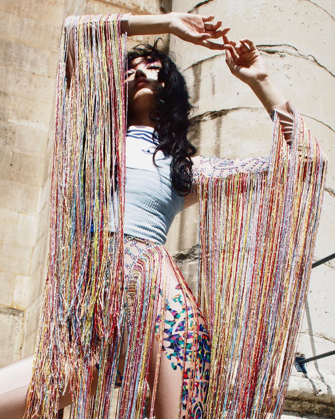 Woman in colorful fringe garment poses against a stone structure, with sunlight highlighting her face and the texture of the garment.