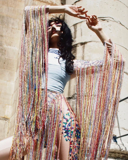 Woman in colorful fringe garment poses against a stone structure, with sunlight highlighting her face and the texture of the garment.