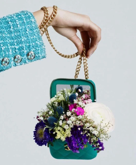 Close-up of a hand holding a quilted turquoise purse filled with colorful flowers and with a chunky gold chain strap on a grey background.