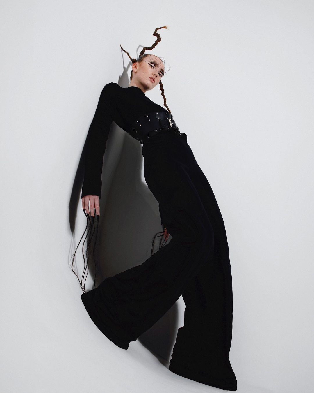 A slender woman in a long-sleeved black top, corset, and bell-bottom trousers poses against a bright white wall in a fashion shoot.
