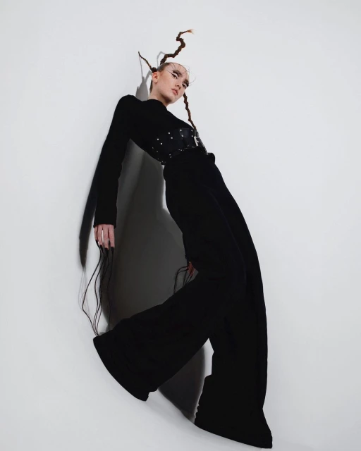 A slender woman in a long-sleeved black top, corset, and bell-bottom trousers poses against a bright white wall in a fashion shoot.