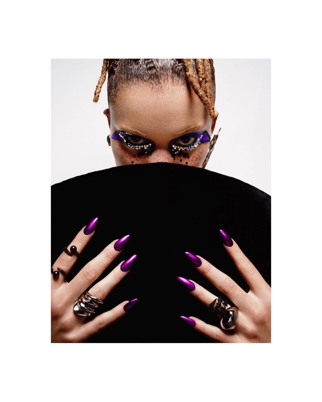 Close-up studio shot of a woman with creative eye makeup, braided hair, and long, pointed, shiny purple nails. She wears statement rings.