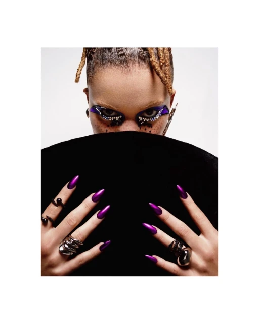 Close-up studio shot of a woman with creative eye makeup, braided hair, and long, pointed, shiny purple nails. She wears statement rings.