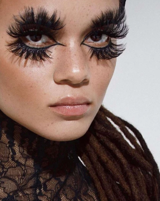 Close-up portrait of a person with dramatically long, feathery eyelashes and bold eyeliner, against a plain white background. Hair is styled into brown dreadlocks.