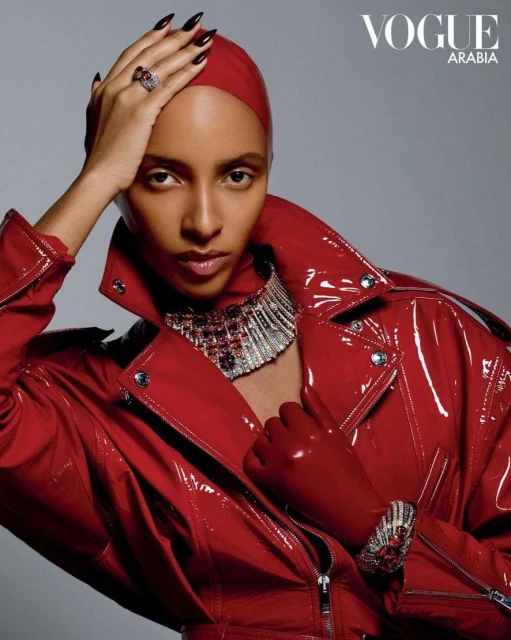 Elegant fashion editorial featuring a woman in a glossy red jacket, matching head covering, gloves, and silver necklace on gray background.