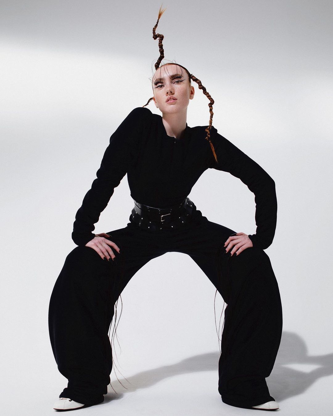 A single fashion model in dramatic black outfit poses in an expressive stance with stylized hair on a bright, clean white backdrop.