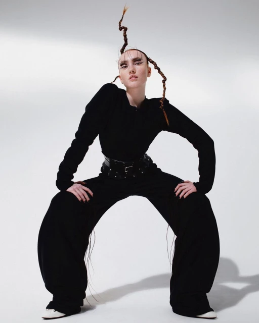 A single fashion model in dramatic black outfit poses in an expressive stance with stylized hair on a bright, clean white backdrop.