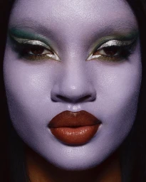 Close-up portrait of a person with purple skin, glossy dark lips, and green and silver eye makeup by Jabe in Paris.