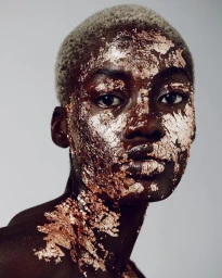 Close-up portrait of a Black model with short dyed hair and metallic gold leaf flaky makeup on face and neck.
