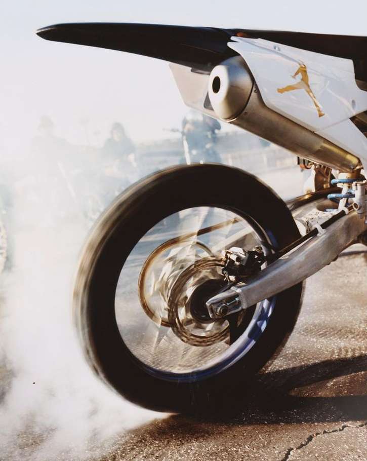 Rear view of a white and black motorbike with a golden logo, tire spinning on pavement creating a cloud of smoke. Outdoor setting.