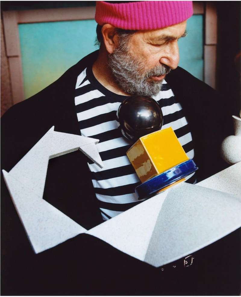 A man with a pink hat and striped shirt gazes at a geometric object in front of a blue and green background. The image has a soft, artful quality.