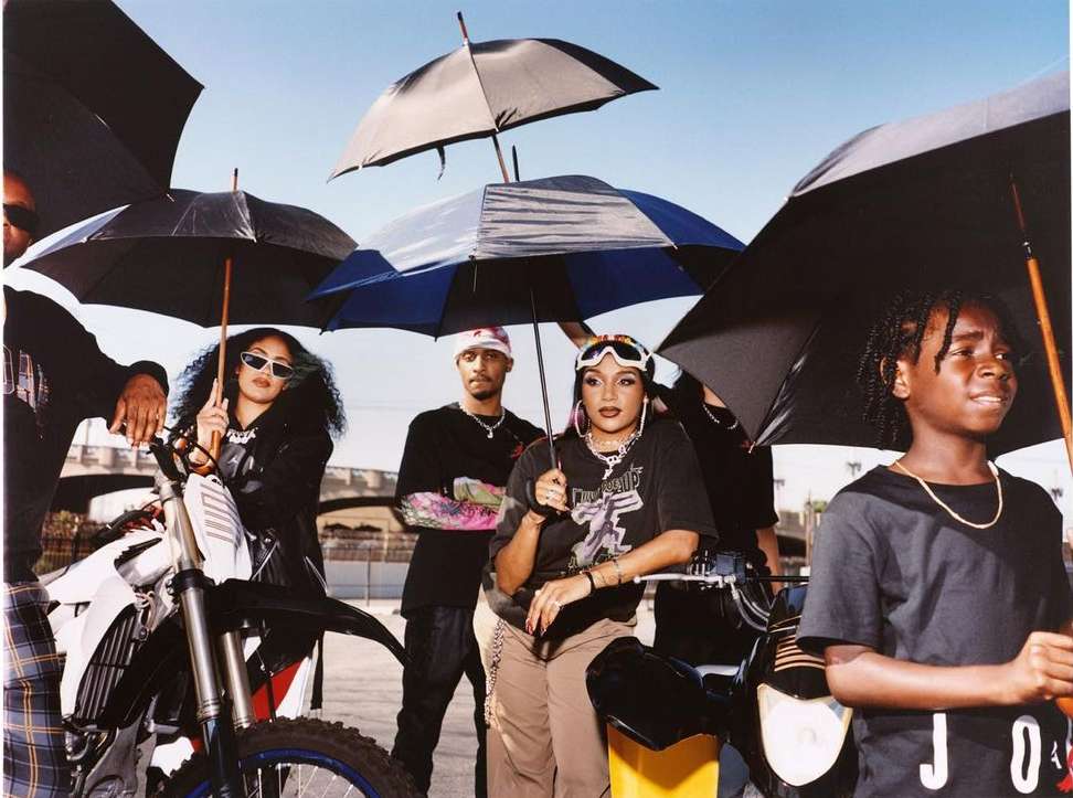 Group of young adults pose with motorcycles under umbrellas in an outdoor urban setting on a bright day, blending fashion and motorsports.
