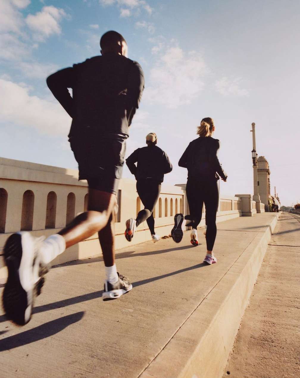 Four people are running on a concrete walkway with a low concrete barrier, on a sunny day with a blue sky and light clouds.