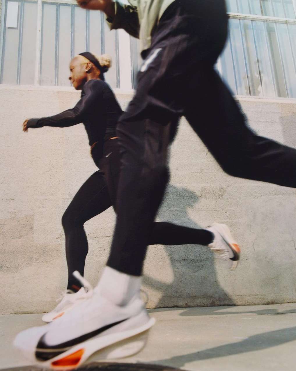 Two adult women in athletic wear run outdoors, showcasing white running shoes with a black swoosh against a light concrete backdrop.
